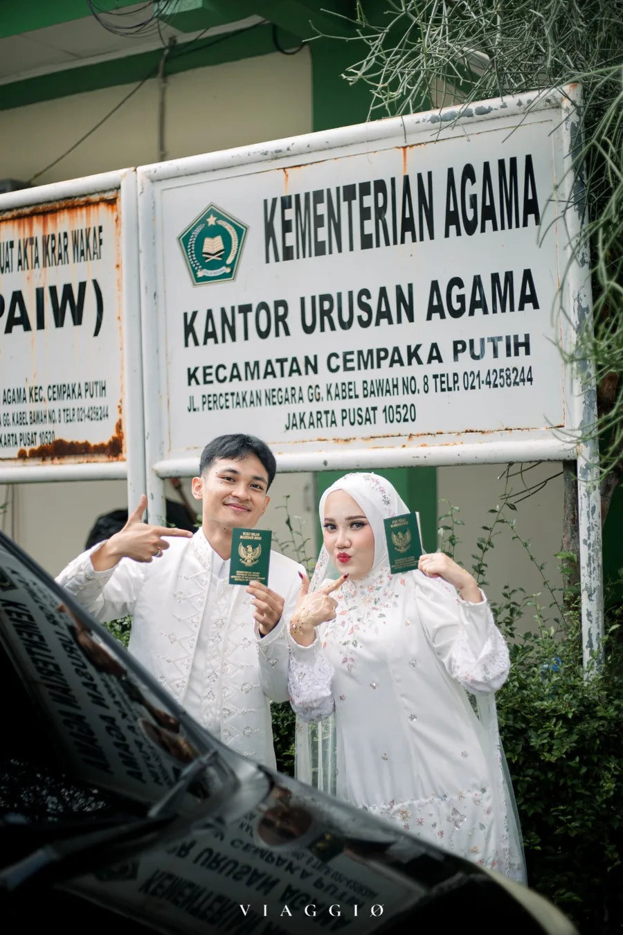 Pasangan dengan buku nikah di depan papan KUA